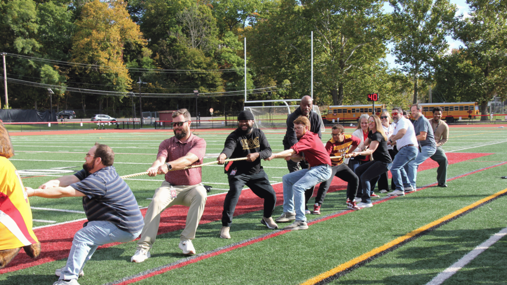 A group of people pull on a rope on a sports field. Trees and parked school buses in background.