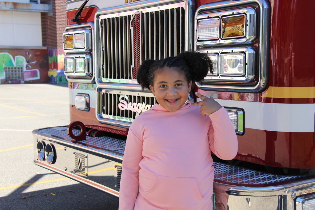 A young girl in a pink shirt stands by a red fire truck, smiling, with buildings and graffiti in the background.