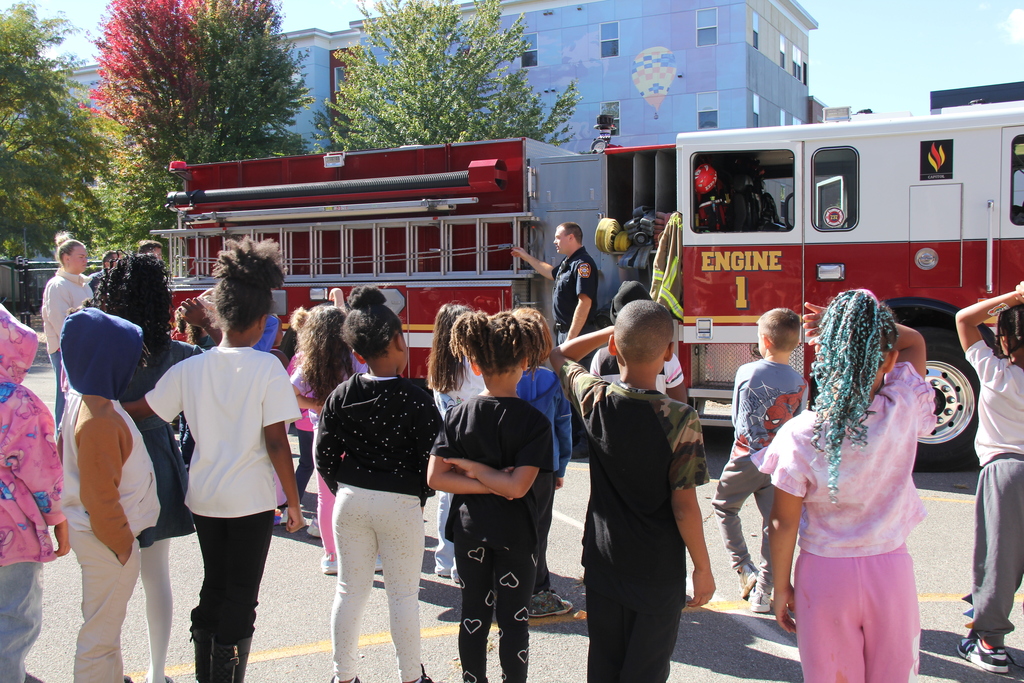 Group of children standing around a red fire truck. Buildings are in the background.