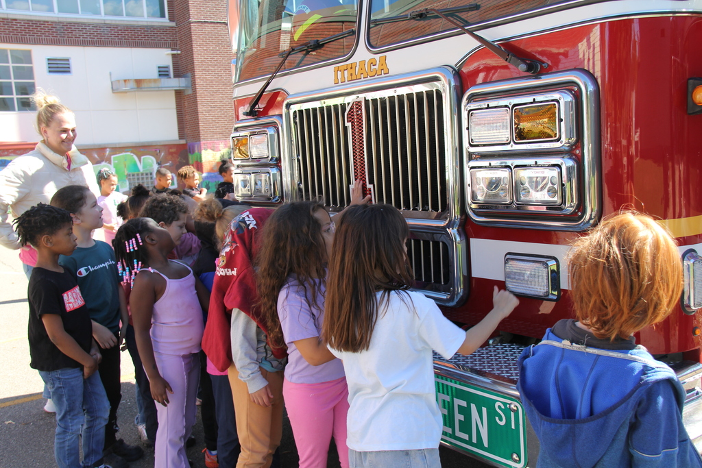 Group of children stand near a red fire truck, examining it closely. A man stands behind them.