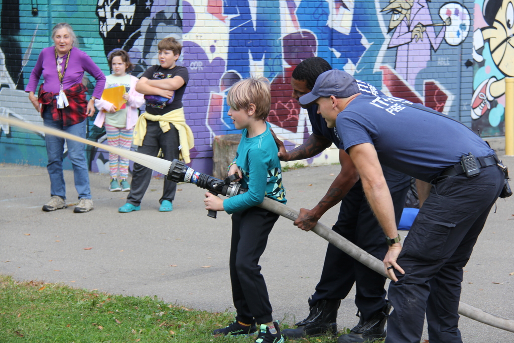A firefighter helps a young child aim a hose outside while others watch; a colorful mural is in the background.