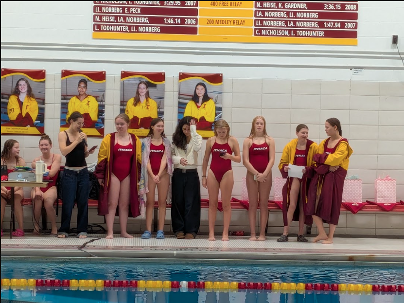 Athletes in swim uniforms stand by a pool, facing sideways. Two women hold papers, one in white and the other in maroon.