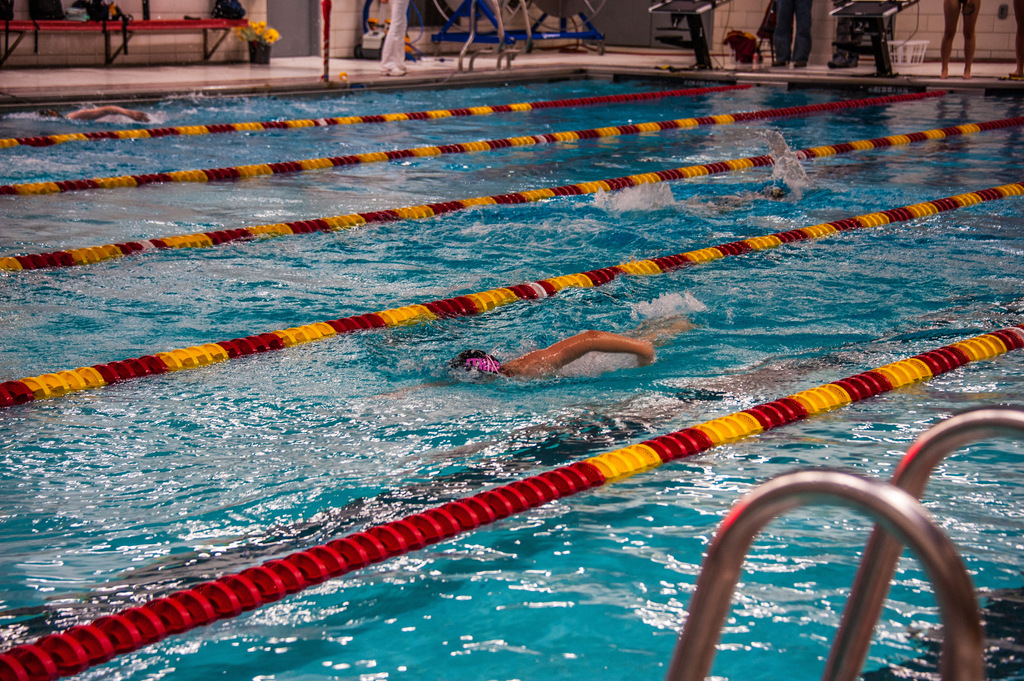 Swimmers in lanes, wearing swim caps and goggles, in an indoor pool with red, yellow, and white lane markers.