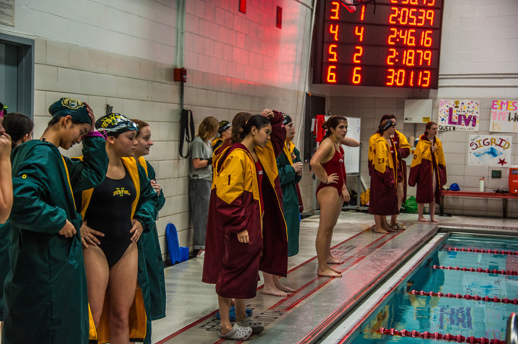 Athletes in swimsuits stand at the edge of a pool. Scoreboard behind them displays times. Some wear robes, others are barefoot.
