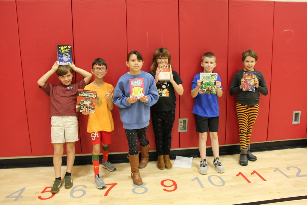Students stand in a row, holding books, next to a red wall with numbers and letters on the floor.