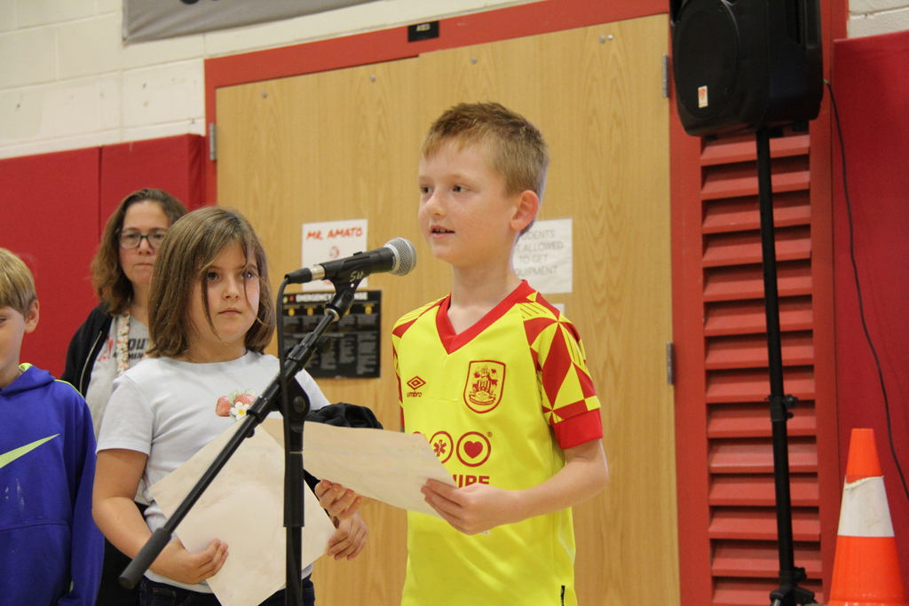 A boy in a yellow and red shirt reads from a piece of paper into a microphone.