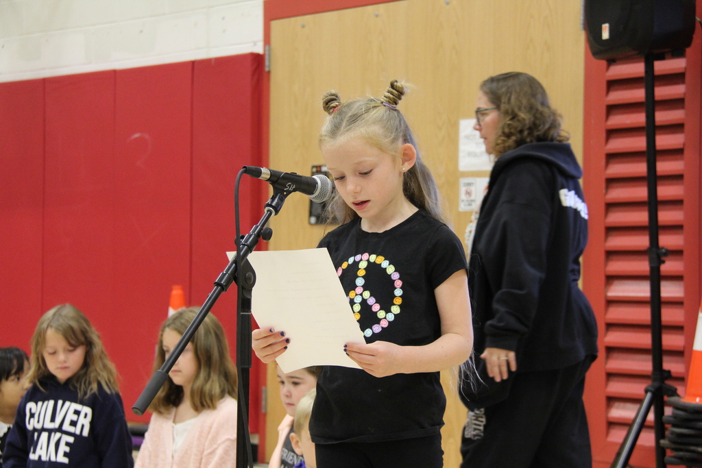 Child standing at microphone, holding paper, with several children sitting behind her and a woman standing.