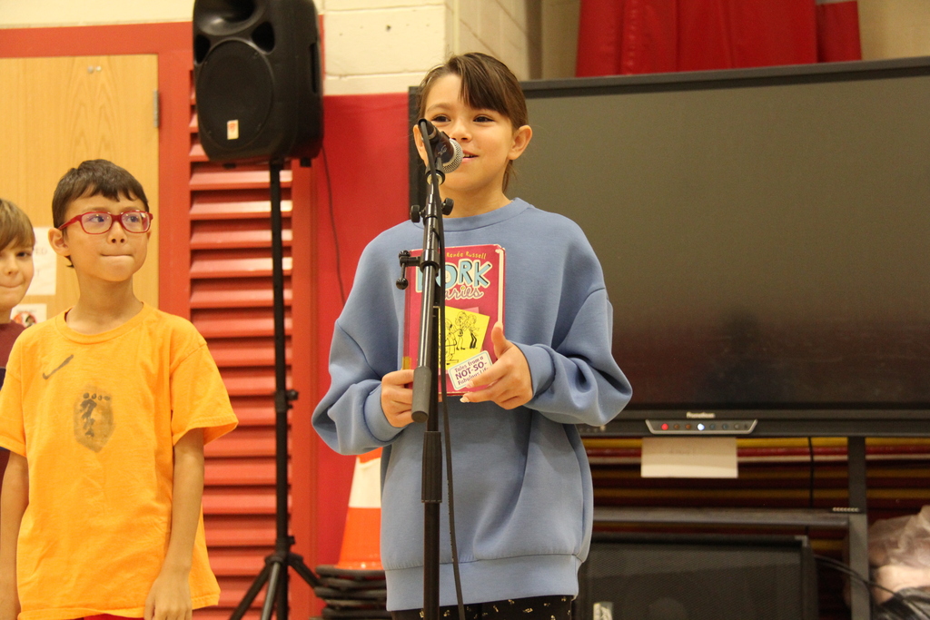 Two students in front of a microphone, one holding a book, near a speaker and a television.
