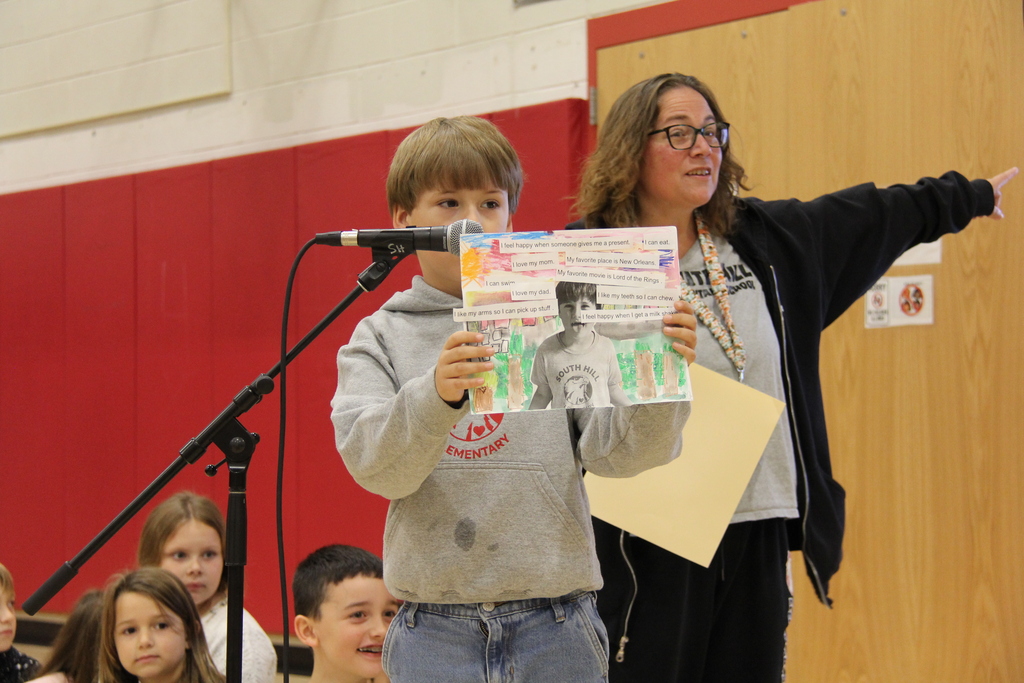 Child holds poster at microphone as adult stands behind; audience of children sit before them.