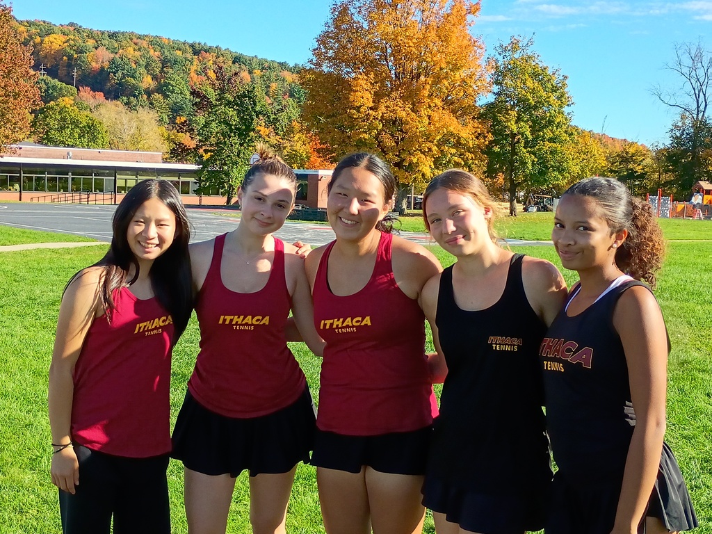 Five young women in athletic wear stand in a row on a green lawn. Behind them are trees and buildings.