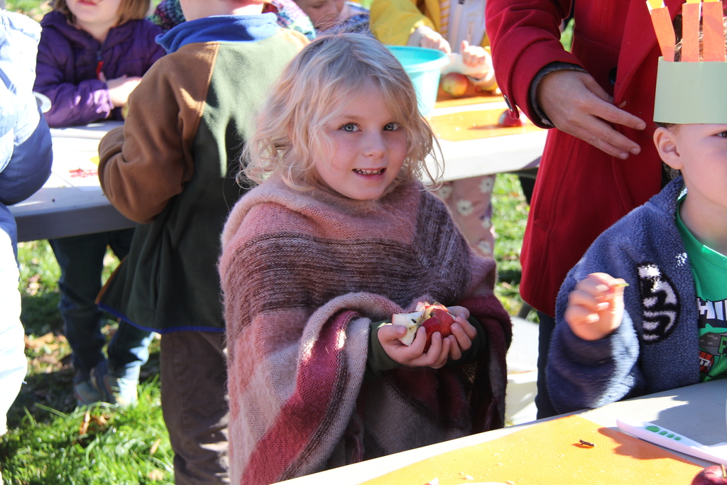 A child in a sweater holds apples in her hand next to a student wearing a paper crown