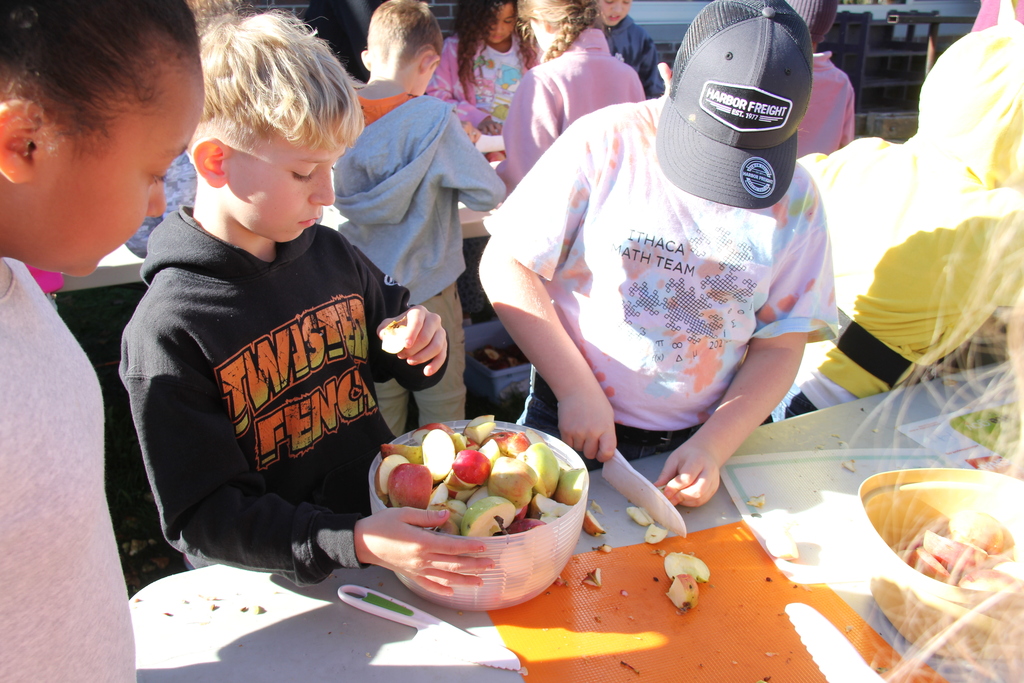 Children prepare food, one holds a bowl of apples, another uses a knife, in an outdoor setting.