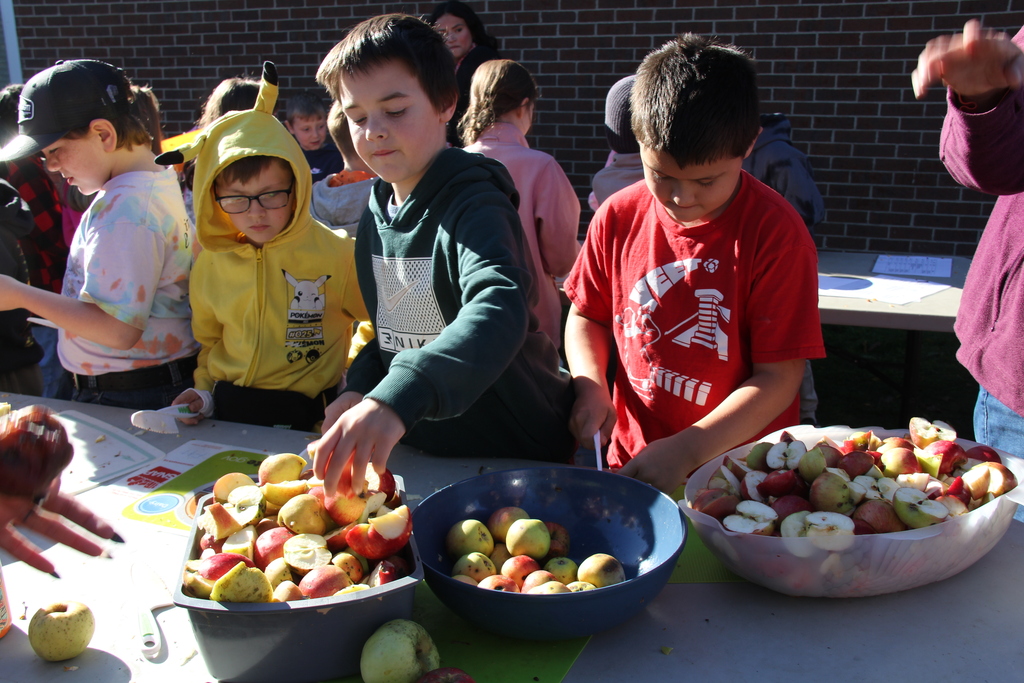 Children around a table with bowls of sliced apples and a boy in a red shirt.