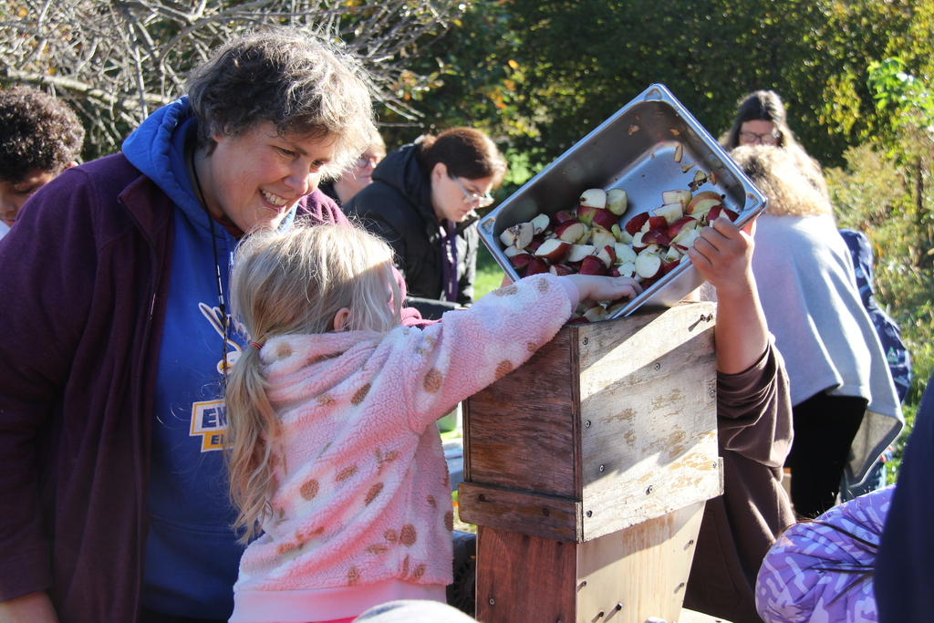 Woman and child holding tray with apple pieces above apple cider press outdoors. Other people are nearby.