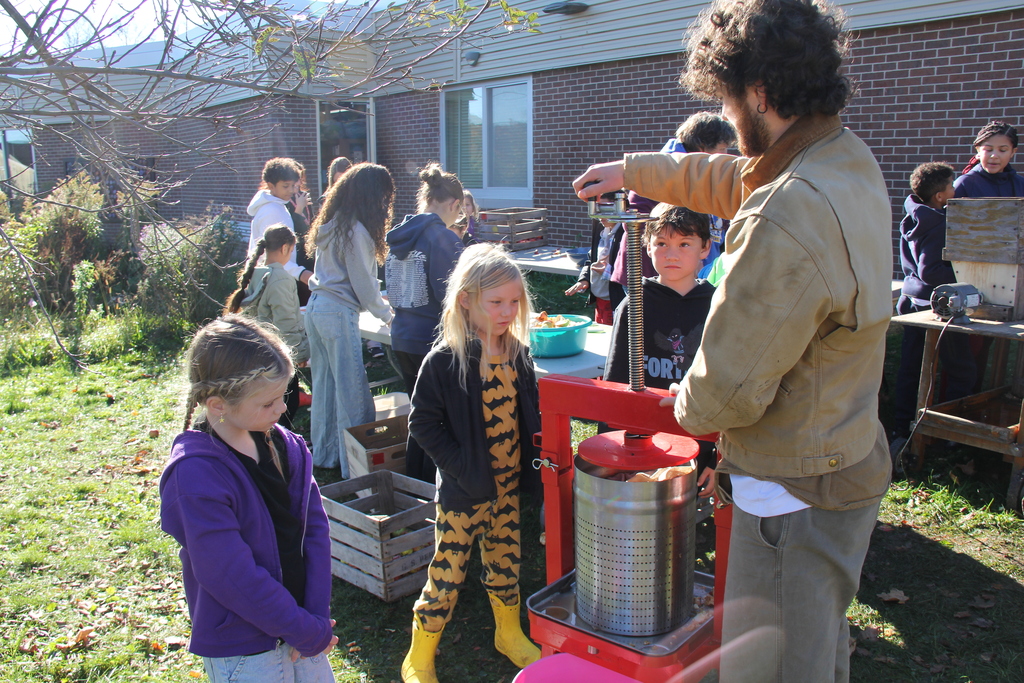 A man demonstrates a fruit press to children, a few of whom hold apples.