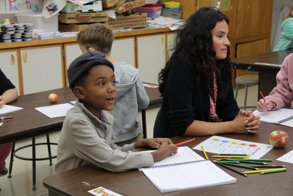 Students seated at desks, one holds a notebook and colored pencils, another holds an apple, while an adult looks on.