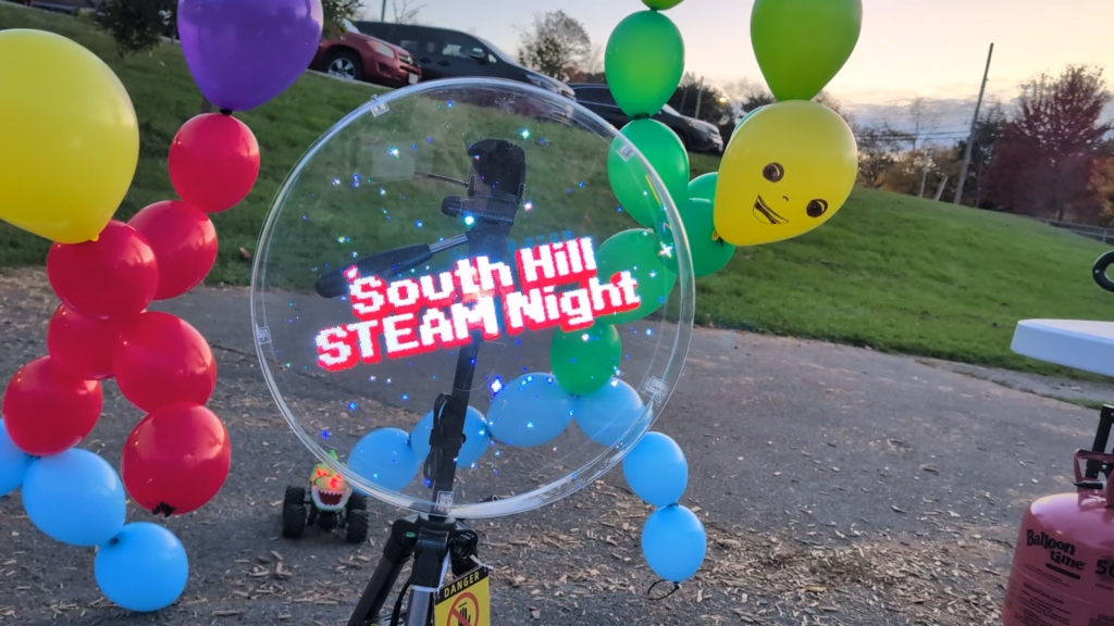 Bright balloons in red, yellow, blue, green, and purple with a smiley face. A glass circle displays the text "SOUTH HILL STEAM NIGHT".