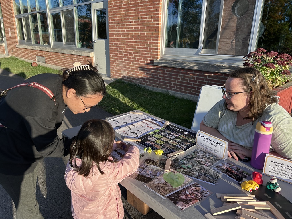 Two adults and a child look at various objects on a table outside a brick building.