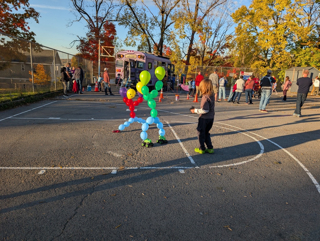 Kids play near balloon figures and a food truck on a basketball court, with trees and a fence in the background.