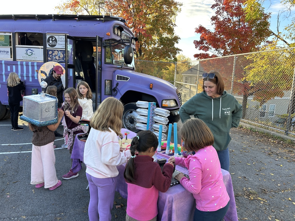 A group of children and a woman are gathered around the physics bus playing with science toys