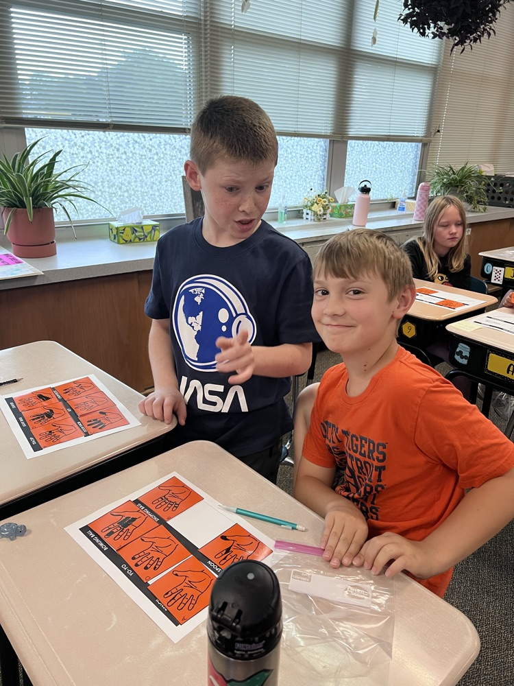 Sixth-grade students at a classroom desk examine inked handprints on paper and compare them to a light bulb, a doorknob, and a pencil.