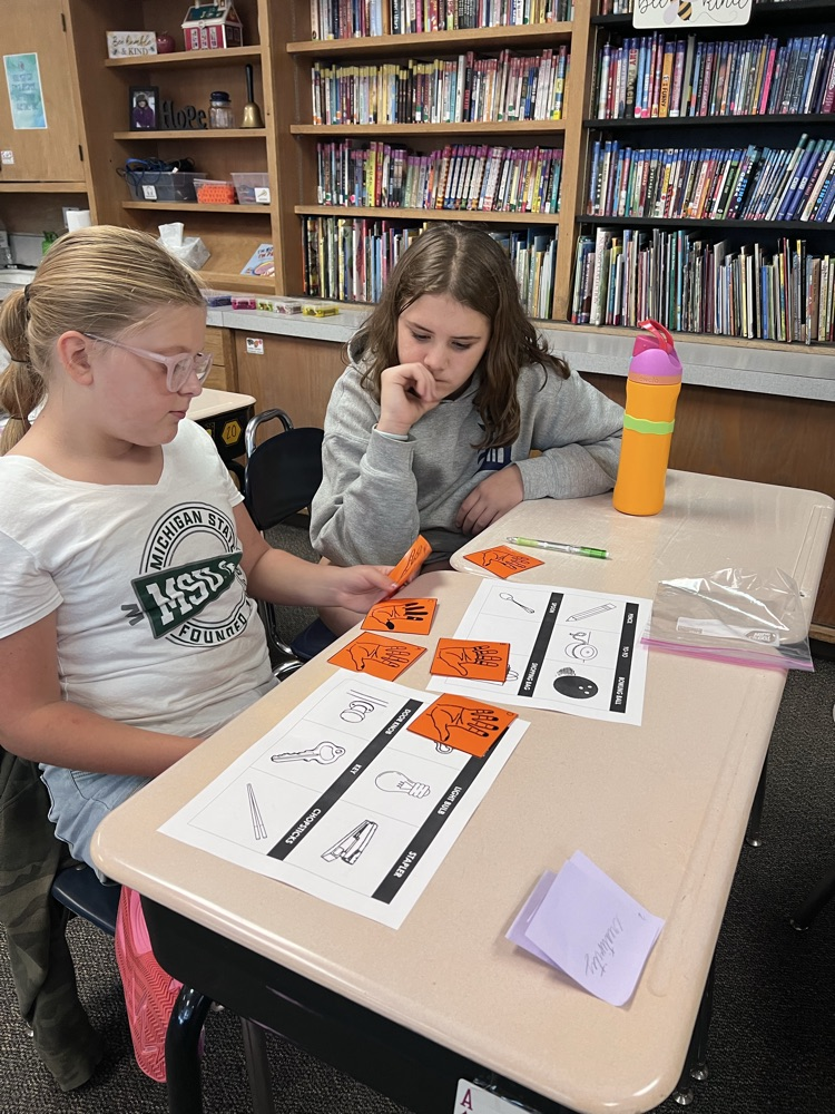 Sixth-grade students at a classroom desk examine inked handprints on paper and compare them to a light bulb, a doorknob, and a pencil.