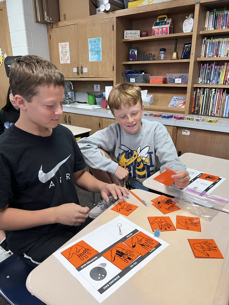 Sixth-grade students at a classroom desk examine inked handprints on paper and compare them to a light bulb, a doorknob, and a pencil.