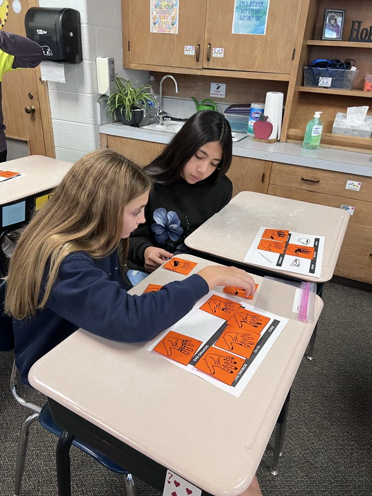 Sixth-grade students at a classroom desk examine inked handprints on paper and compare them to a light bulb, a doorknob, and a pencil.