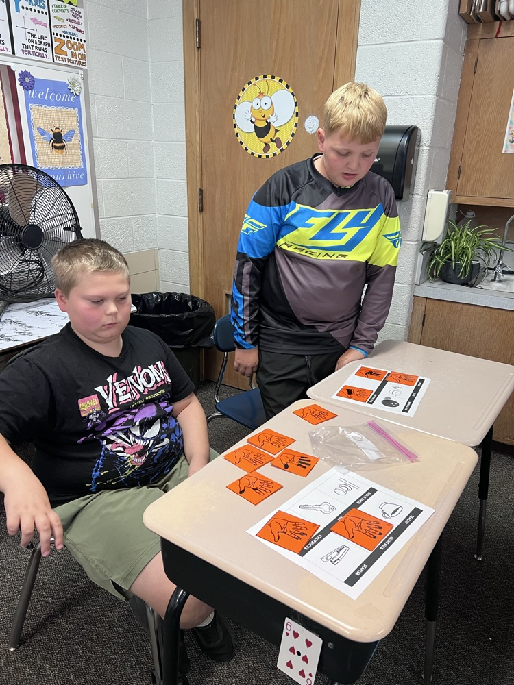 Sixth-grade students at a classroom desk examine inked handprints on paper and compare them to a light bulb, a doorknob, and a pencil.