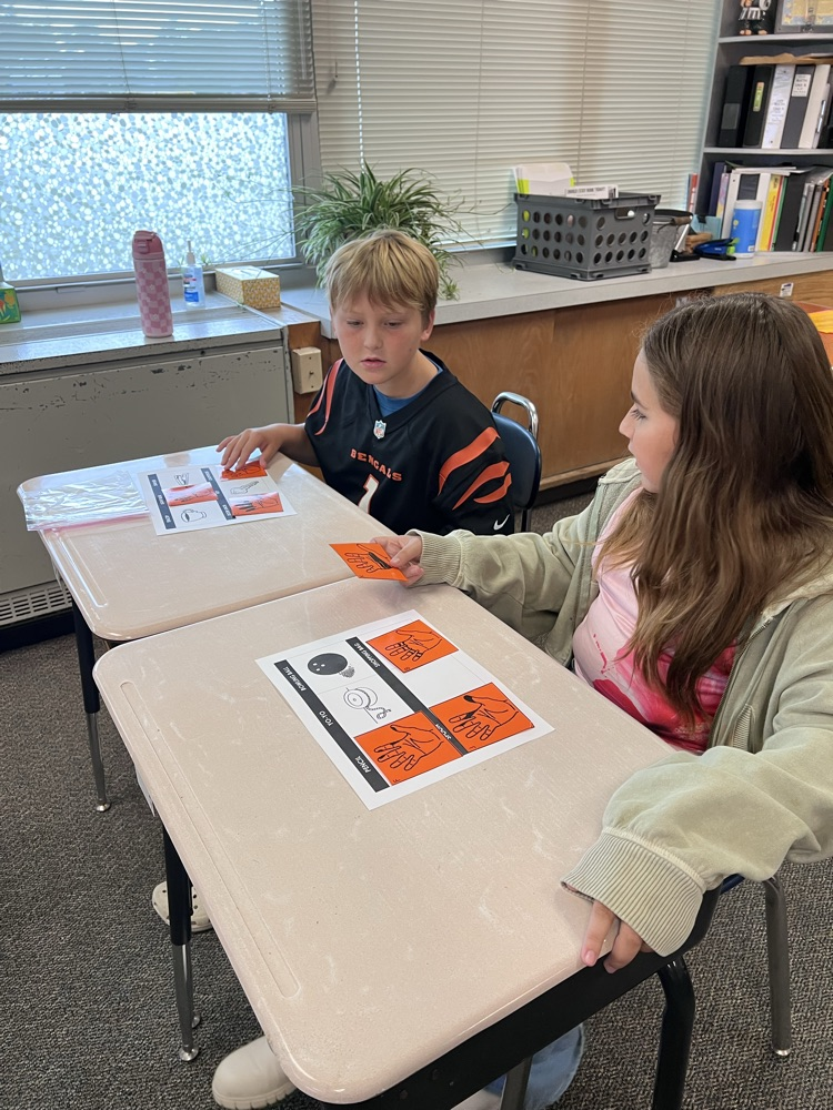 Sixth-grade students at a classroom desk examine inked handprints on paper and compare them to a light bulb, a doorknob, and a pencil.
