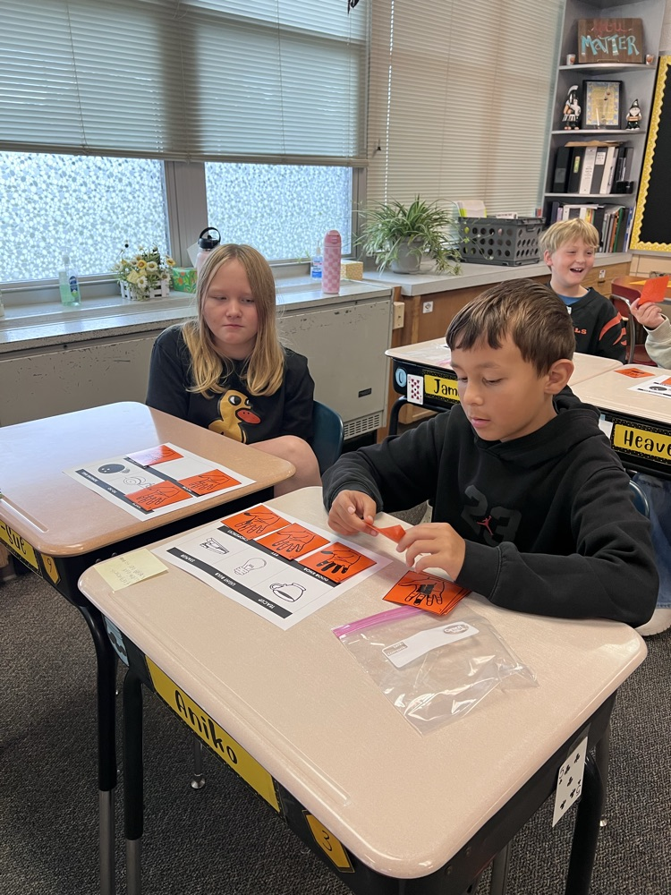 Sixth-grade students at a classroom desk examine inked handprints on paper and compare them to a light bulb, a doorknob, and a pencil.