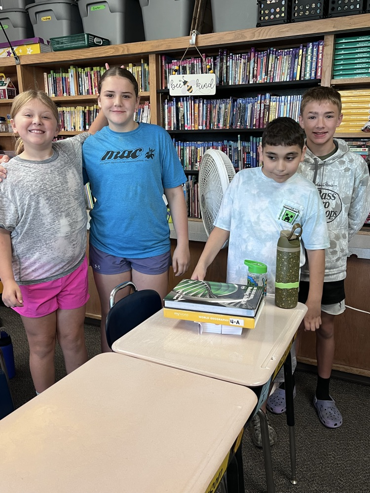 Four 6th grade students standing behind a desk, presenting a bridge built from folded index cards that successfully holds up two heavy textbooks in a classroom setting.