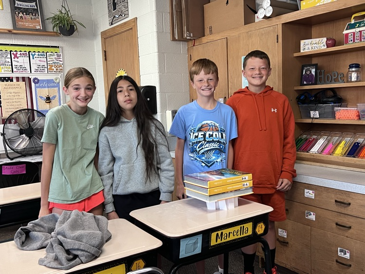 Four 6th grade students standing behind a desk, presenting a bridge built from folded index cards that successfully holds up two heavy textbooks in a classroom setting.