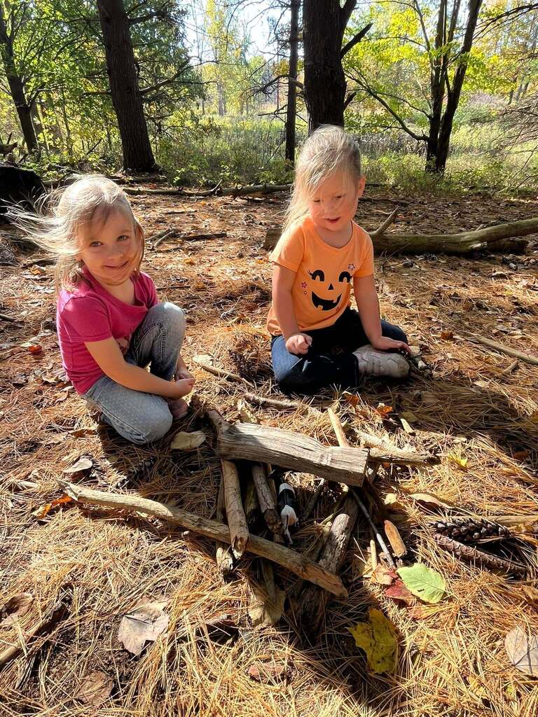 Two students sit on the ground near a pile of sticks.