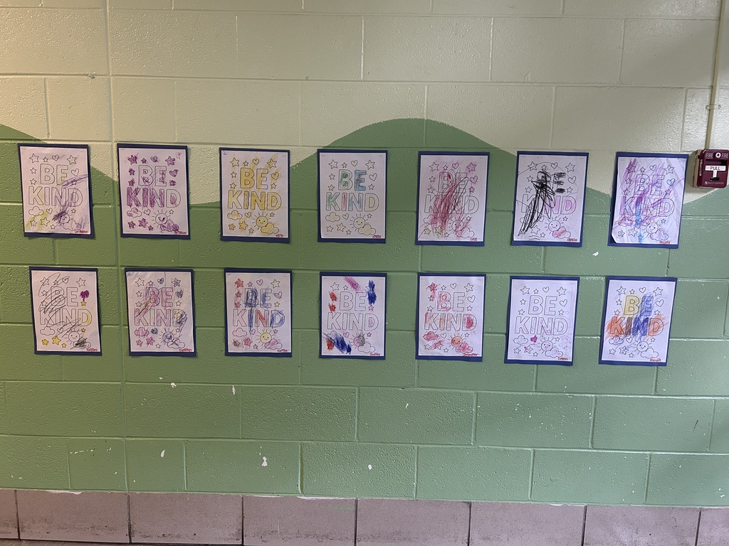 A hallway display at Homer Adkins Pre-K Center features colorful “BE KIND” artwork created by Pre-K 3 scholars. The drawings are displayed in two rows against a green and cream painted brick wall, showcasing each scholar’s unique coloring and creativity in celebration of Kindness Day.