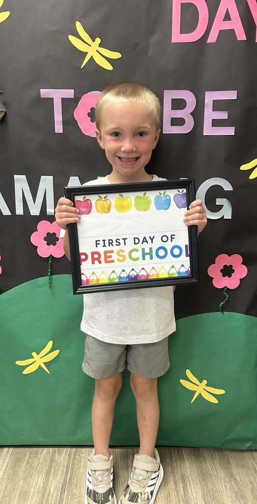 Child smiling holds sign reading "FIRST DAY OF PRESCHOOL" in front of a colorful backdrop.
