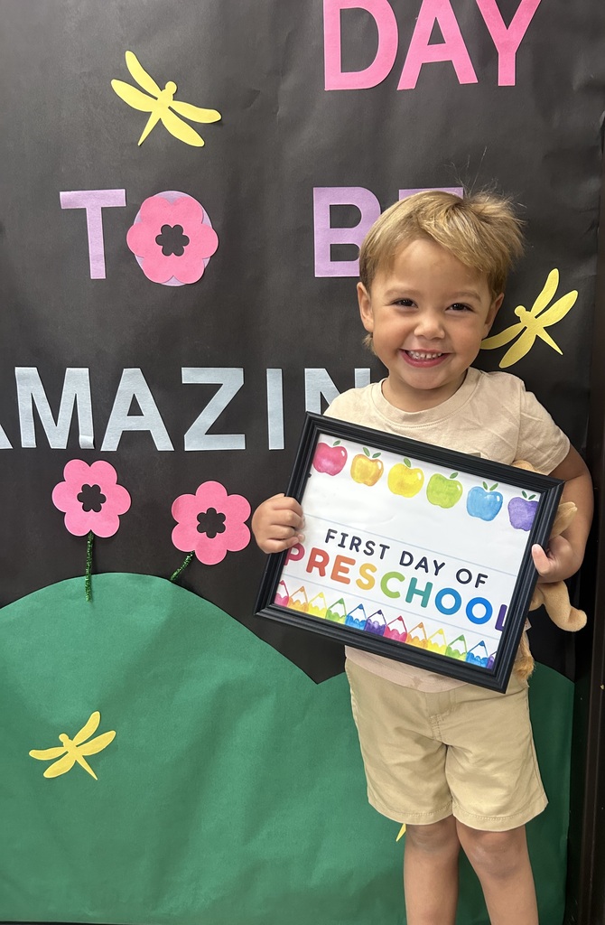 Young child wearing shorts, smiling, holds a framed certificate. Black backdrop has pink and yellow letters. Green hill and flowers.