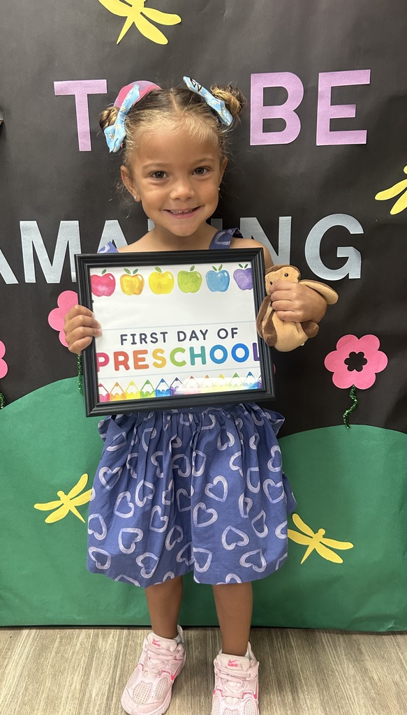 Child holds a framed certificate for first day of preschool, standing against a colorful backdrop with letters and flowers.