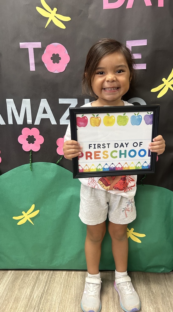 A smiling child holding a framed certificate with "First Day of Preschool" in front of a green backdrop.