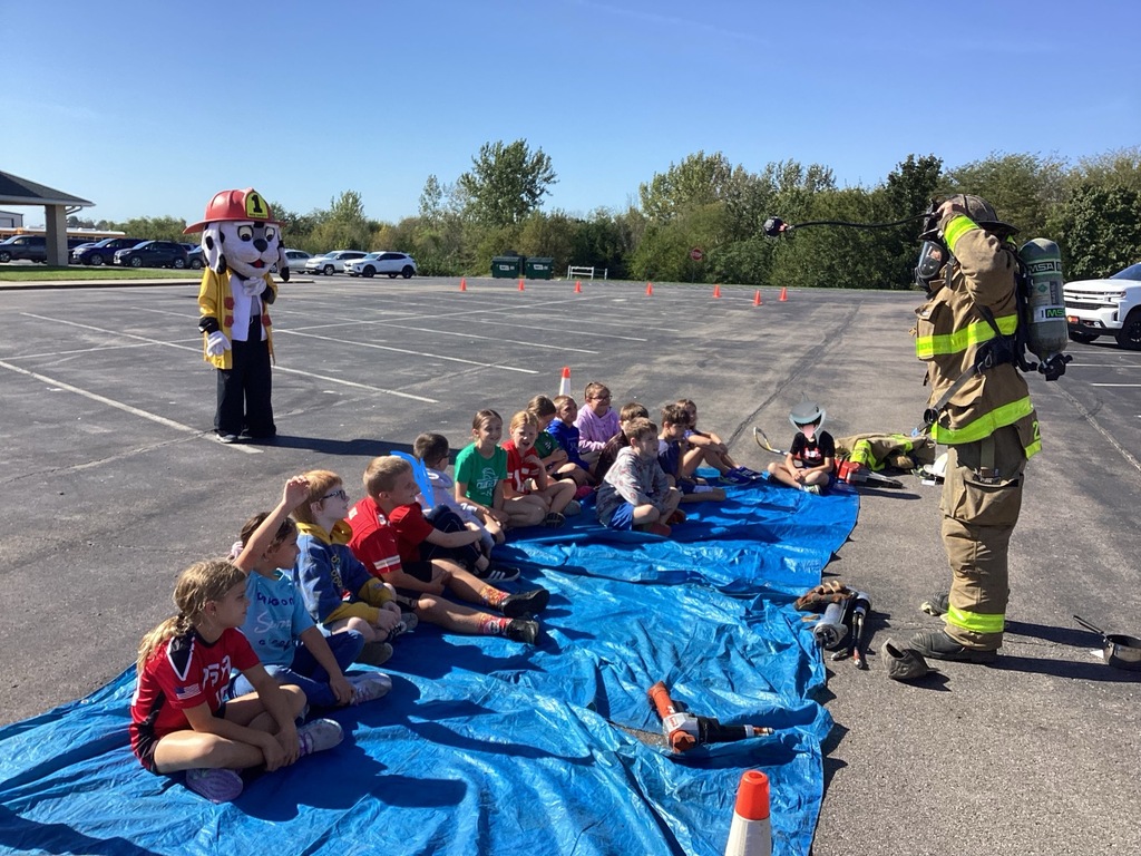 Students sit on ground listening to firefighter.