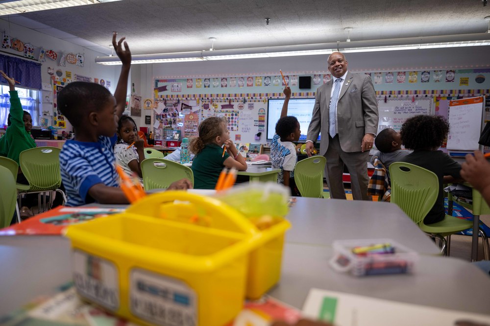 Dr. Brian Yearwood in a suit stands in a brightly decorated classroom, looking at a diverse group of young students with their hands enthusiastically raised.