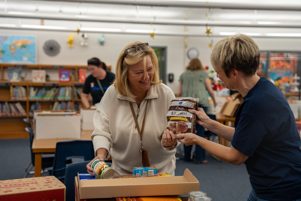 A woman handing another woman a bag of brown sugar and they're both smiling while one of the women is putting a can in a box