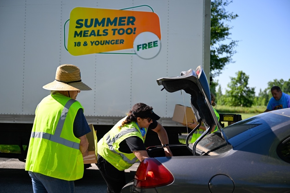 People loading a trunk with food under a sign that says "Summer Meals Too"