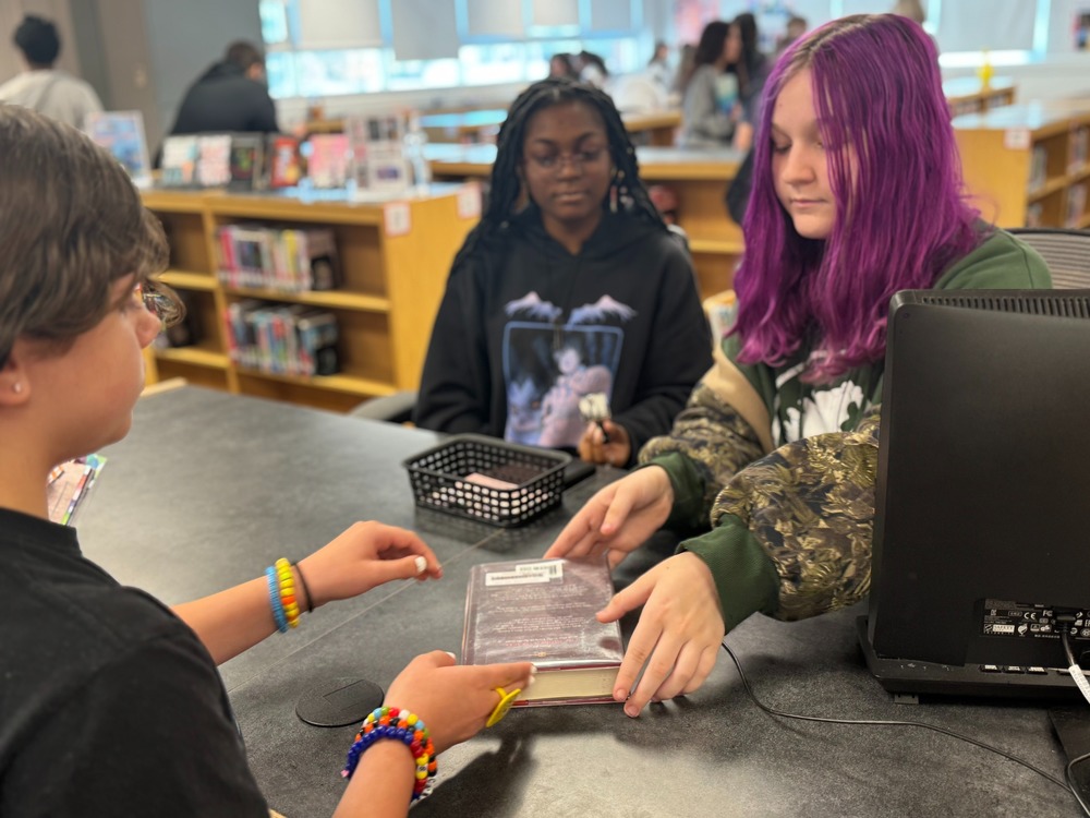Two students working at the PRP High Library helping a fellow student check out a library book.
