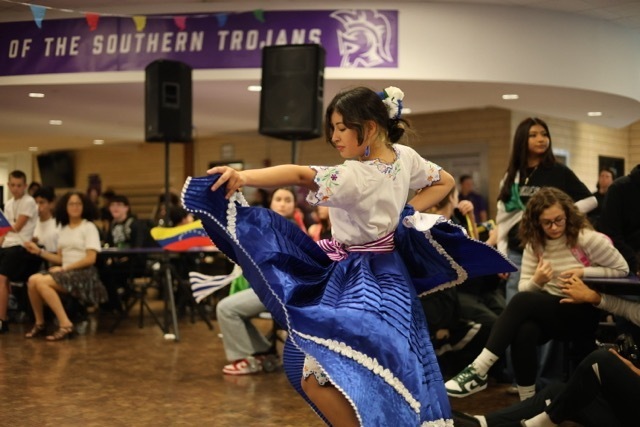 A young woman, seen from the side and back, performs a dance in a school setting. She wears a white embroidered blouse and a large blue skirt with a pink sash.