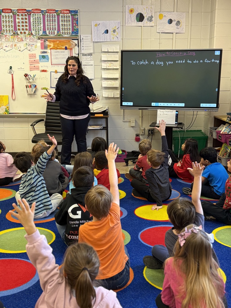 A photograph of a classroom scene.  Foreground: Several elementary-aged students are sitting on a large blue rug decorated with bright, colorful circles. Many students have their hands raised.  Background: A female teacher in a dark sweater stands at the front of the room next to a large digital whiteboard.  Digital Whiteboard: The screen displays the handwritten title "How to Catch a Dog" and the sentence "To catch a dog you need to do a few things".