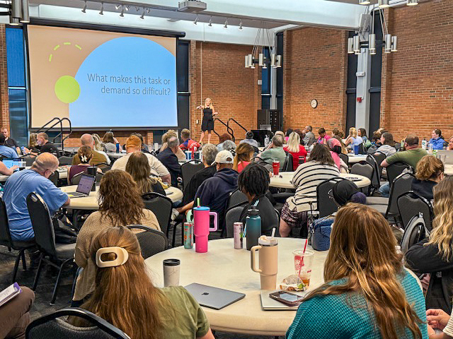 Adults sit at tables while a presenter presents onstage.