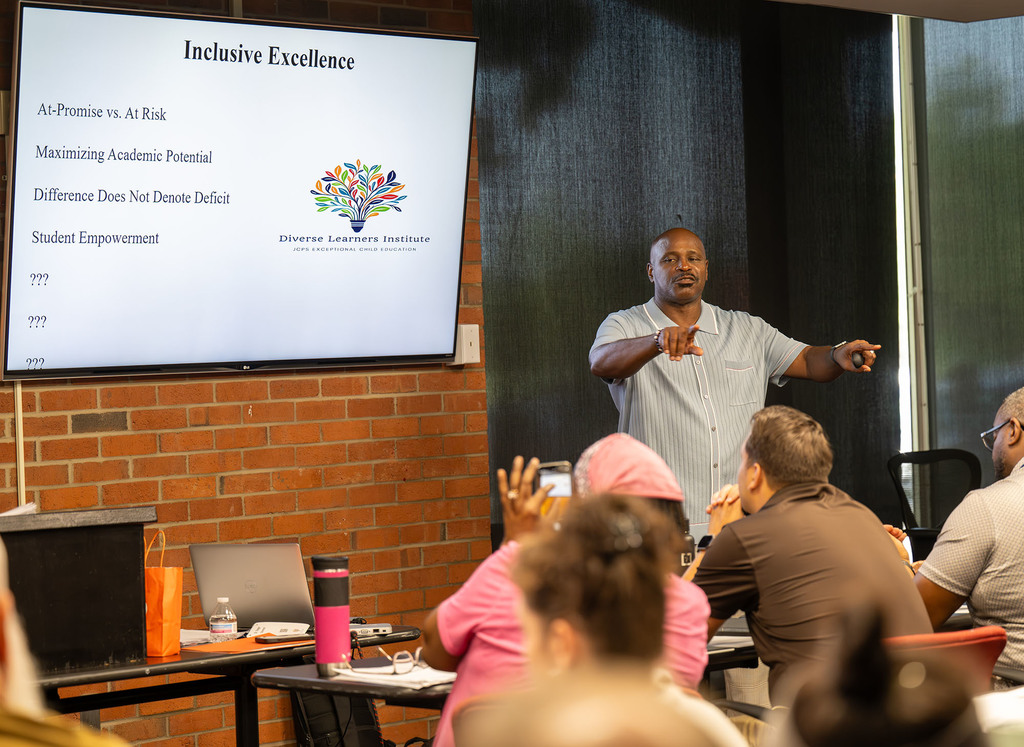 A screen says "Inclusive Excellence, At-Promise vs. At-Risk, Maximizing Academic Potential, Difference Does Not Denote Deficit, Student Empowerment" as a session leader points at participants.
