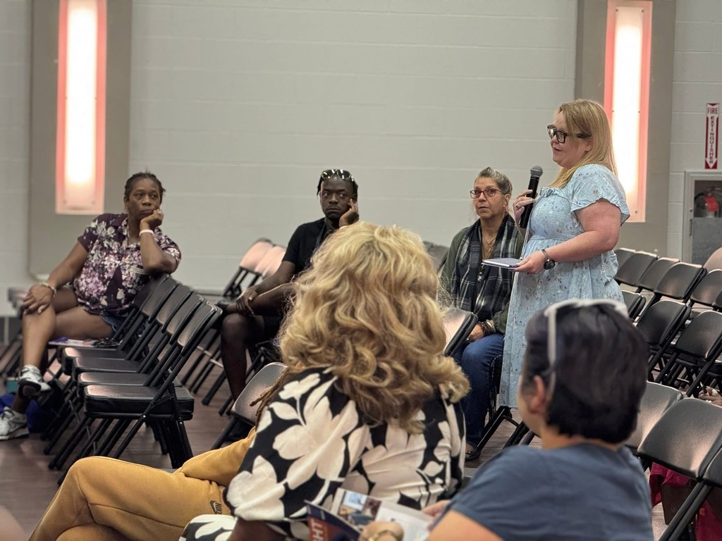 Community members sit in chairs. One stands and speaks into a microphhone. 