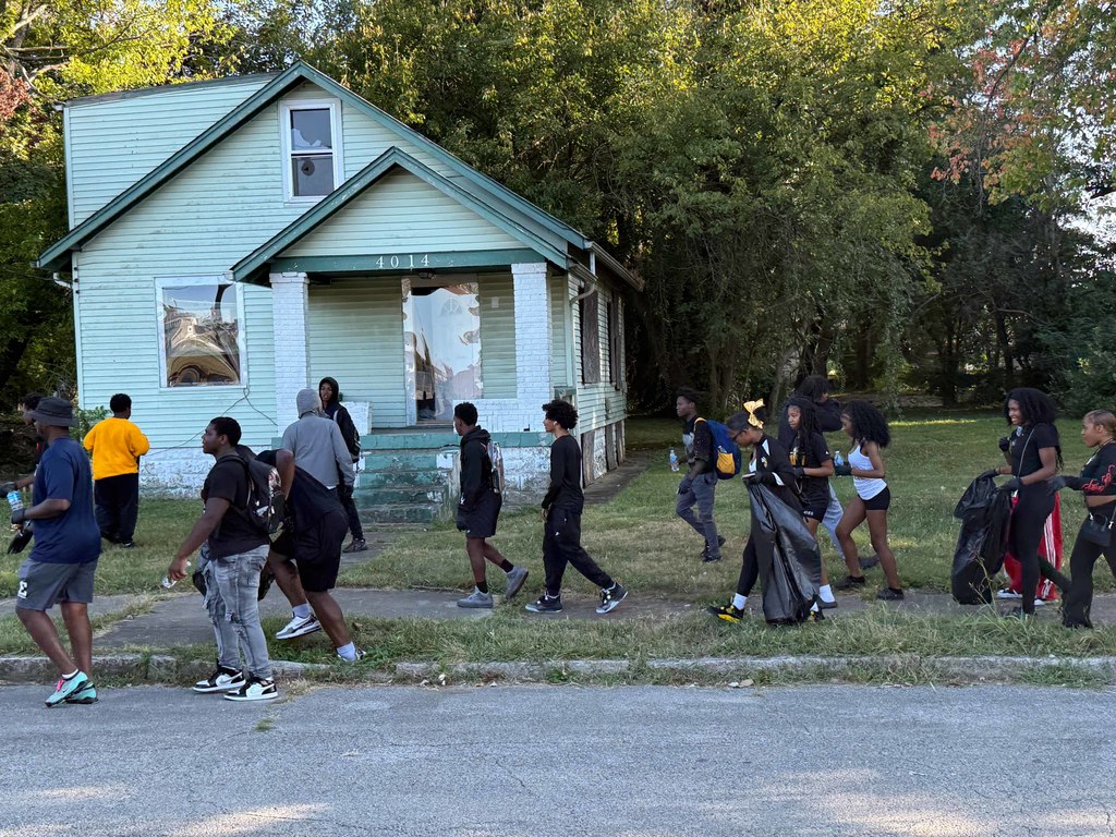 Students walk down a sidewalk and street with a house in the background.